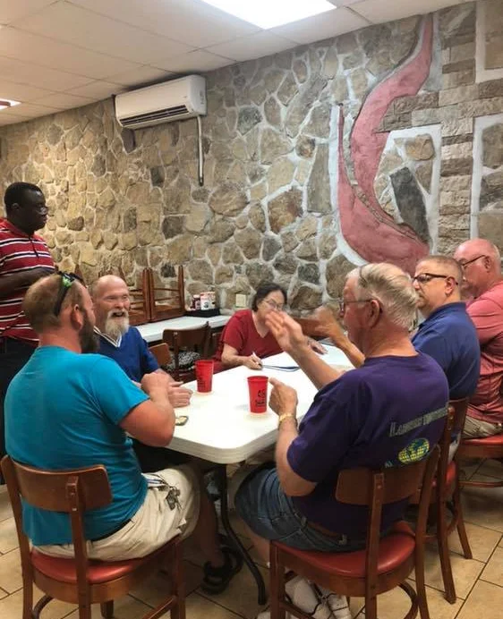 Seven deaf people converse around a table in a casual café with stone walls and a decorative stone mural with the Methodist logo on it. Cups and papers are on the table.