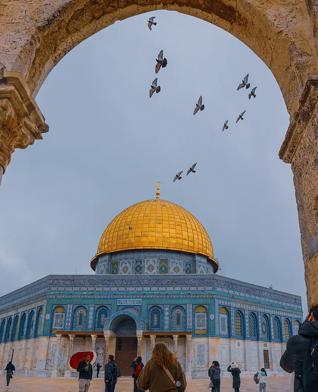 People walk under an arch toward the Dome of the Rock, with birds flying overhead and a cloudy sky in the background.