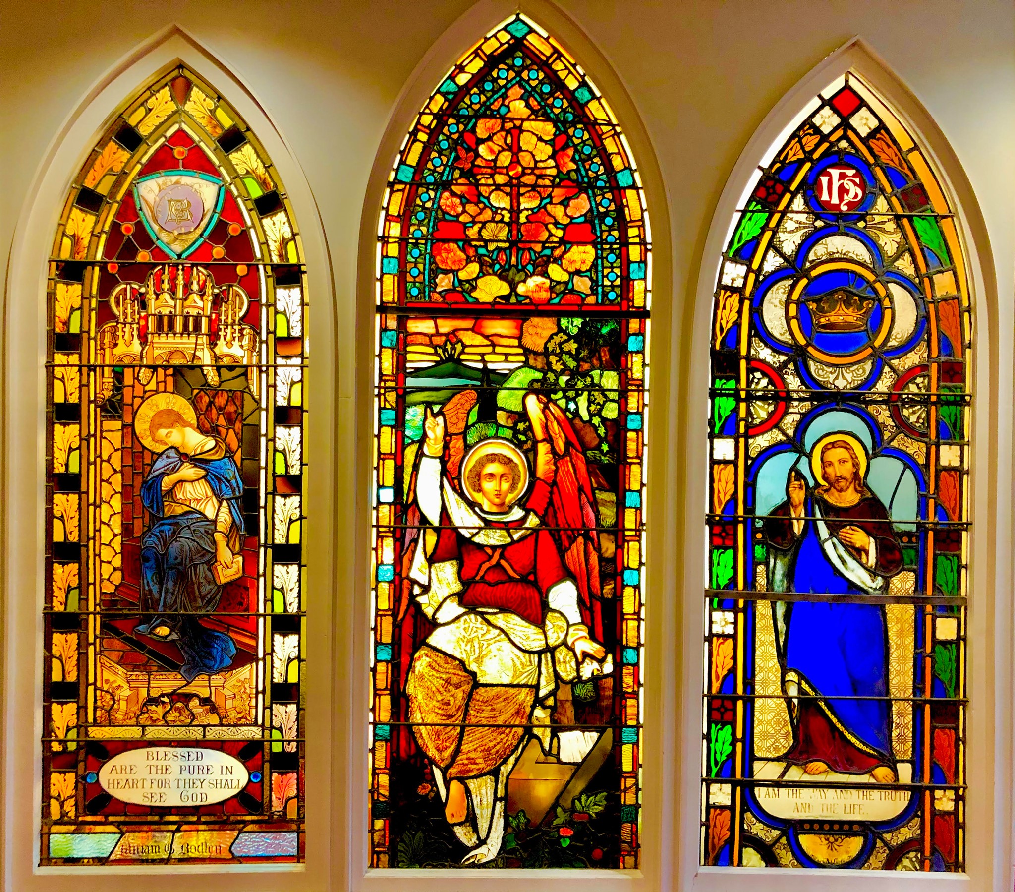 Three vibrant stained glass windows depict religious scenes and figures, including angels and a haloed person, with intricate patterns and vivid colors.